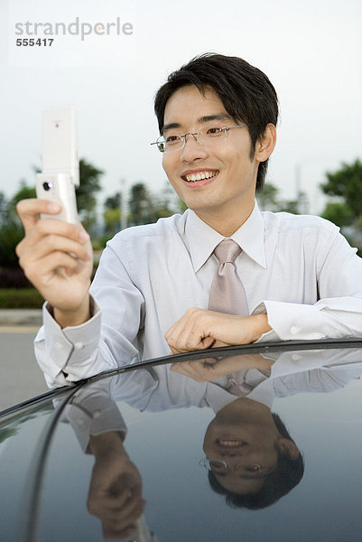 Junger Geschäftsmann lehnt sich an das Auto  hält Handy hoch  lächelt