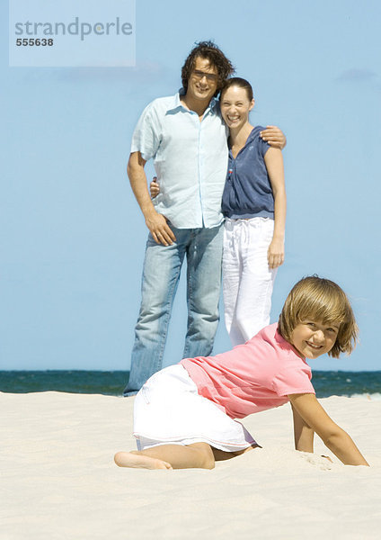 Mädchen graben im Sand  während die Eltern im Hintergrund stehen.