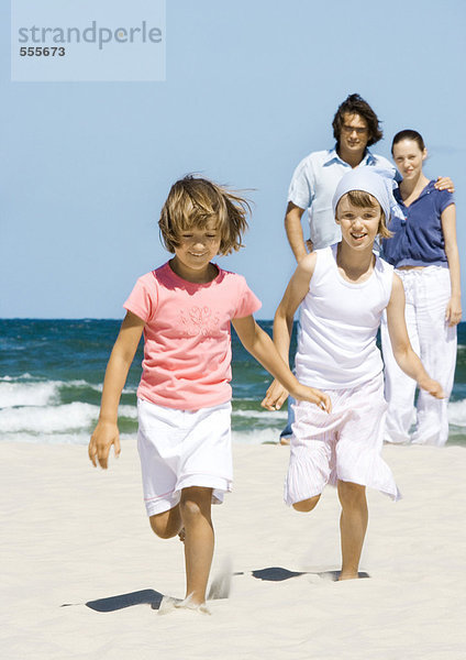 Mädchen laufen am Strand  während die Eltern im Hintergrund stehen.