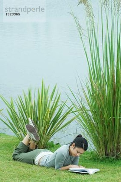 Junge Frau  die auf Gras in der Nähe eines Gewässers liegt und liest.