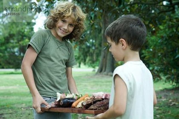 Zwei Jungen halten ein Tablett mit gegrilltem Fleisch.