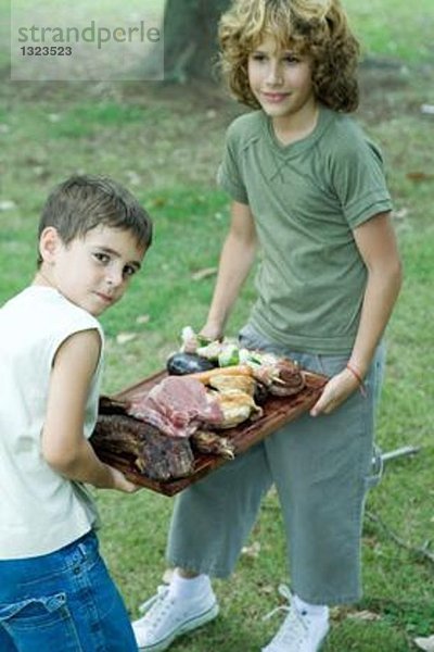 Zwei Jungen tragen ein Tablett mit gegrilltem und rohem Fleisch.