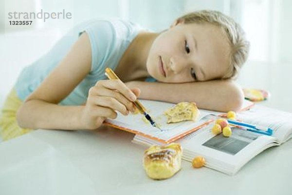 Mädchen am Tisch sitzend  Hausaufgaben machend  Kopf auf dem Arm liegend  kritzelnd  Essen auf Büchern verstreut