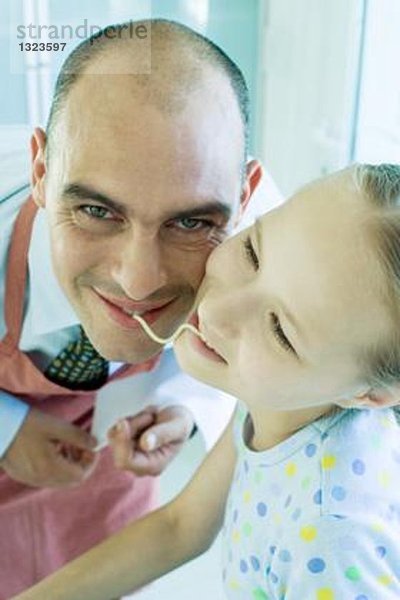 Vater und Tochter halten Enden von Nudeln im Mund  lächelnd
