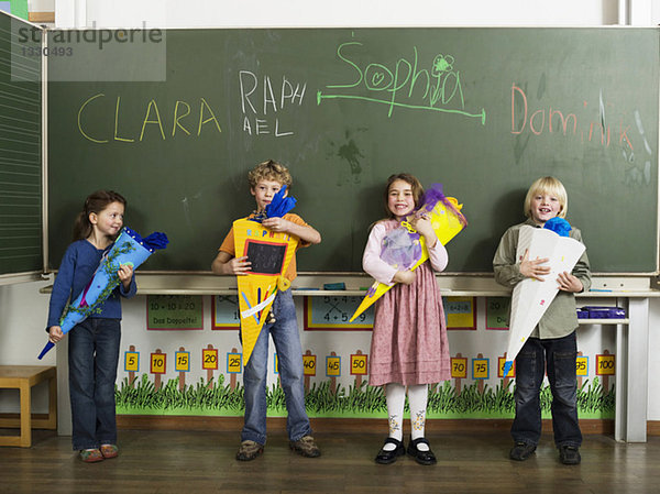 Kinder (4-7) stehen vor der Tafel und halten Schulkegel.