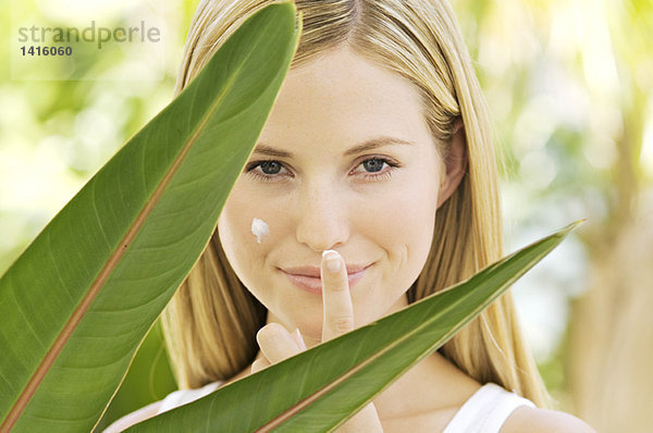 Porträt einer jungen Frau  die in die Kamera schaut  Feuchtigkeitscreme auf ihr Gesicht aufträgt  Zimmerpflanzenblätter  im Freien