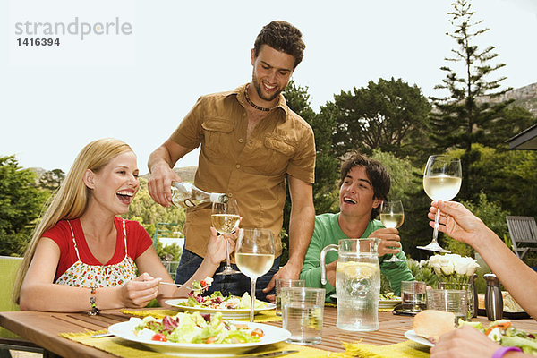 2 junge lächelnde Paare sitzen am Gartentisch  Mann schüttet Wein aus