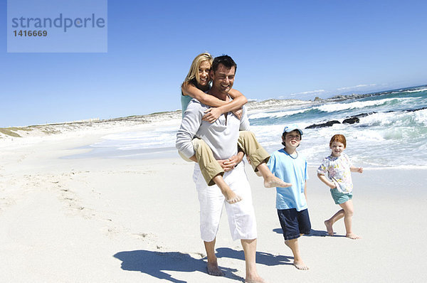Eltern und zwei Kinder laufen am Strand  Mann trägt Frau auf dem Rücken  im Freien