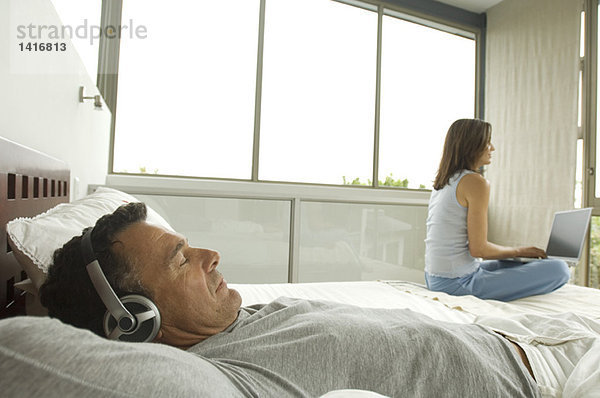 Paar im Schlafzimmer  Mann hört Musik  Frau nutzt Laptop  drinnen