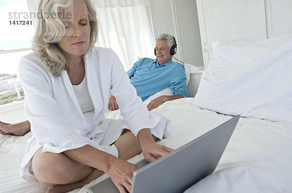Frau mit Laptop im Schlafzimmer  Mann mit Kopfhörer im Hintergrund
