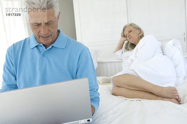 Paar im Schlafzimmer  Mann mit Laptop  Frau im Hintergrund liegend