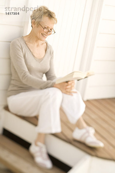 Lächelnde Seniorin beim Lesen eines Buches  auf der Holzterrasse sitzend