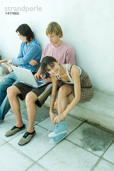 Gruppe junger Freunde auf der Bank sitzend  eine junge Frau bindet Schuhbänder und lächelt in die Kamera.