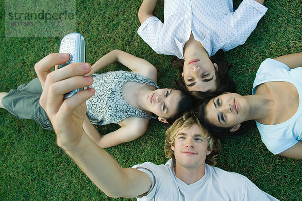 Gruppe junger Freunde auf Gras liegend  Kopf zusammen  ein junger Mann beim Fotografieren mit dem Handy