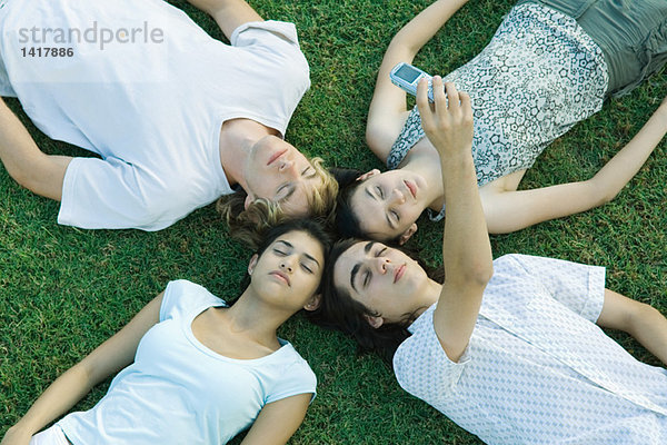 Gruppe junger Freunde  die mit geschlossenen Augen auf Gras liegen und mit dem Handy fotografieren.