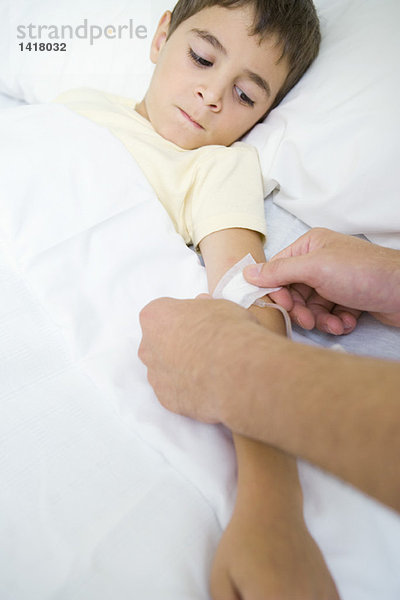 Junge liegt im Krankenhausbett und beobachtet Mann legt Verband über Infusionsschlauch