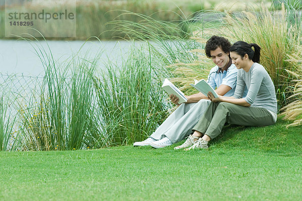 Junges erwachsenes Paar beim Lesen im Freien  sitzend auf Gras am Ufer des Sees