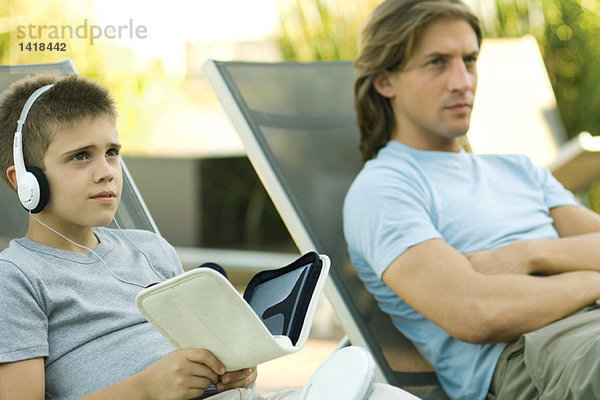 Vater und Sohn sitzen zusammen auf Liegestühlen  der Junge hört Kopfhörer.