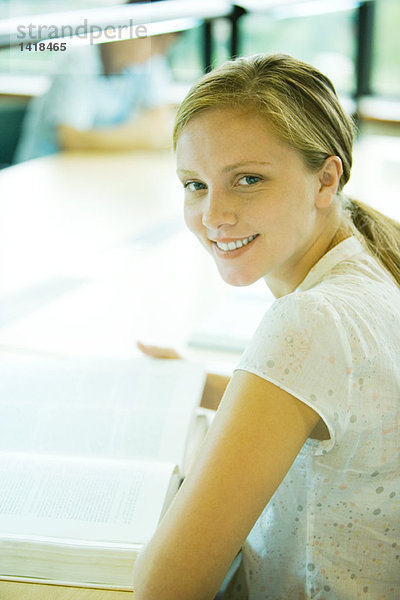 Eine Studentin sitzt am Tisch in der Bibliothek und lächelt in die Kamera.