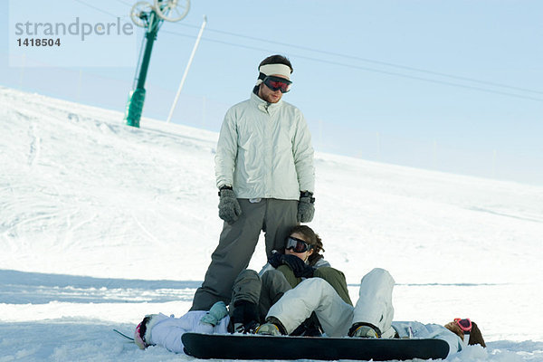 Snowboarder im Schnee liegend  zu einem Freund aufblickend