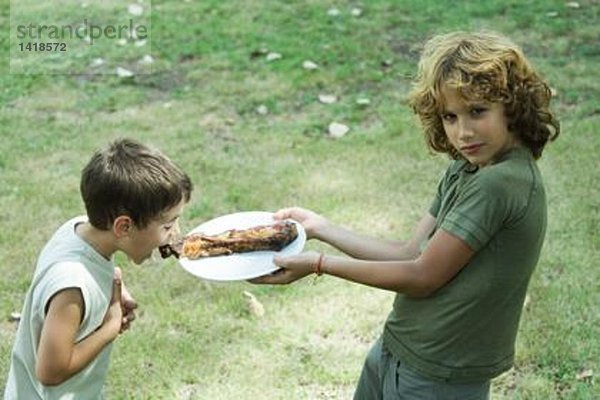 Junge hält den Teller mit gegrilltem Fleisch hoch  während der zweite Junge sich nach vorne beugt  um zu beißen.