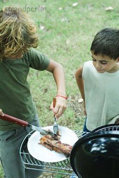 Zwei Jungs stehen neben dem Grill  einer zerlegt Fleisch auf dem Teller.