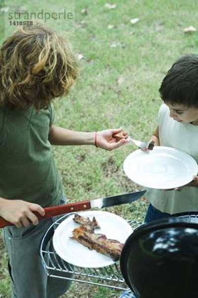 Zwei Jungs stehen neben dem Grill  einer legt ein Stück Fleisch auf den Teller des anderen.