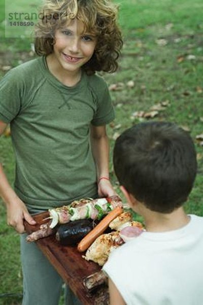 Zwei Jungen tragen ein Tablett mit gegrilltem Fleisch.
