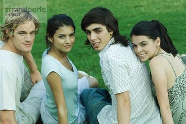 Vier junge Freunde sitzen auf Gras  Seite an Seite  schauen über die Schultern in die Kamera.