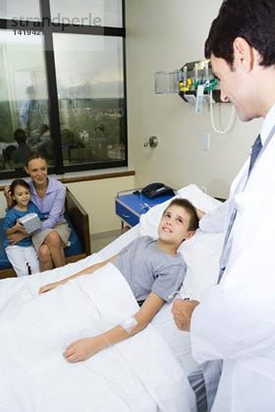 Junge im Krankenhausbett liegend  umgeben von Familie und Arzt