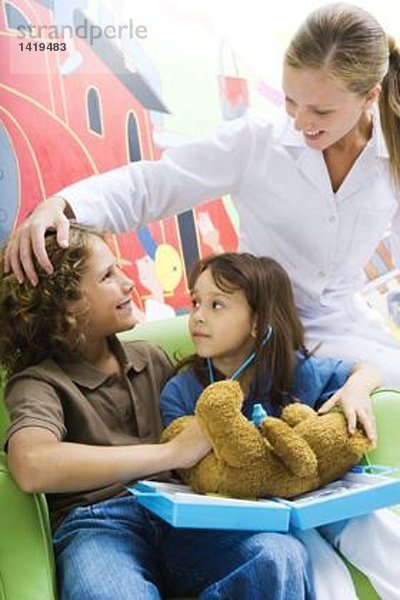 Junge und Mädchen spielen Doktor mit Teddybär  während die Krankenschwester zuschaut.