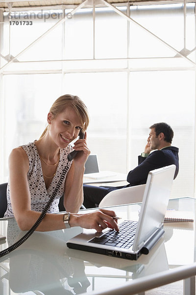 Geschäftsfrau mit Laptop beim Telefonieren  männlicher Kollege im Hintergrund