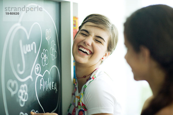 Junge Freundinnen schreiben Namen in Herzen auf Kreidetafel  lachend