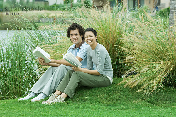 Paar auf Gras sitzend mit Büchern  lächelnd vor der Kamera