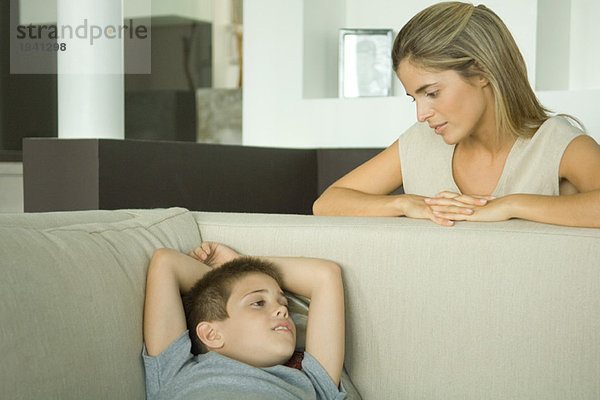 Junge liegt auf dem Sofa und redet mit Mutter  die auf ihn herabblickt.