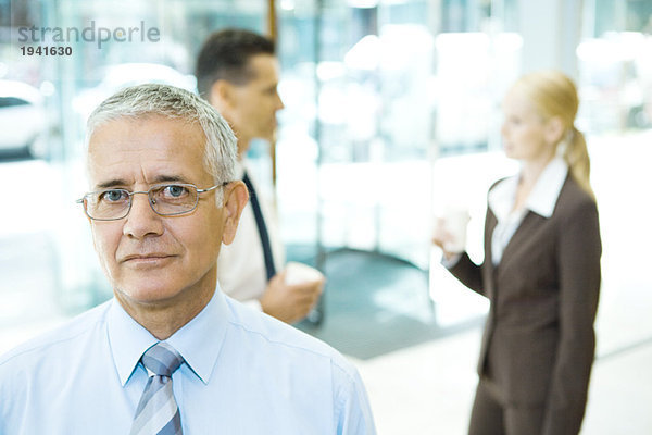 Erwachsener Geschäftsmann mit Blick auf Kamera  Portrait  Mitarbeiter im Hintergrund