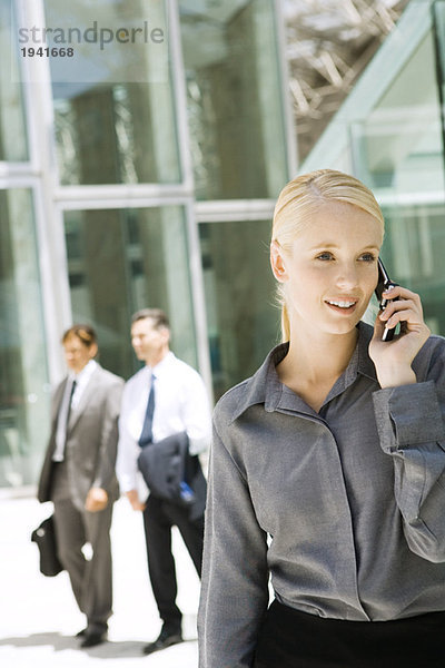 Junge Geschäftsfrau  die draußen mit dem Handy telefoniert  schaut weg  Geschäftsleute  die im Hintergrund laufen