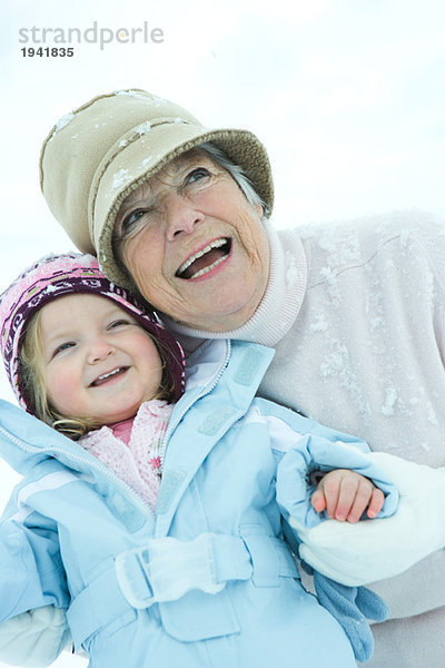 Seniorin umarmt Enkelin im Schnee  beide lächelnd  Taille oben