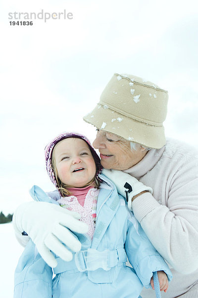 Großmutter umarmt Kleinkind im Schnee  beide lächelnd