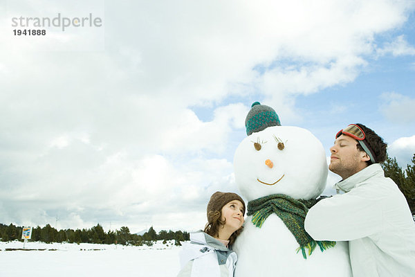 Bruder und Schwester lehnen sich an den Schneemann  beide lächelnd  die Augen des Bruders geschlossen.