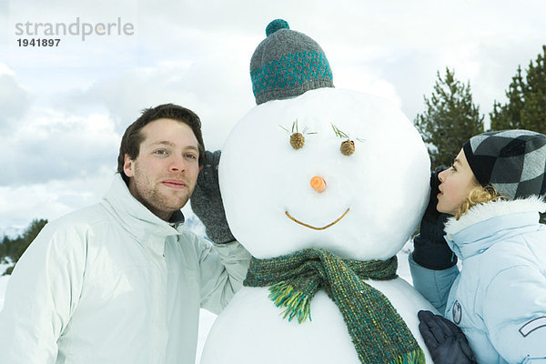 Bruder und Schwester lehnen sich an den Schneemann  Schwester flüstert  Bruder lauscht