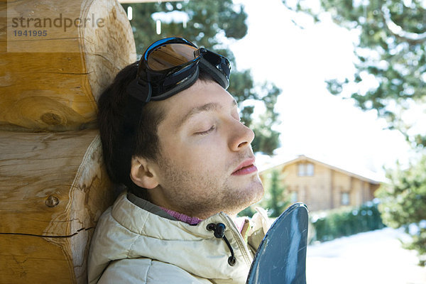 Junger Mann an Blockhütte gelehnt  mit Snowboard  Augen geschlossen