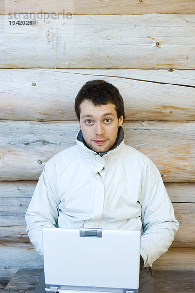 Junger Mann sitzend neben der Blockhauswand  mit Laptop
