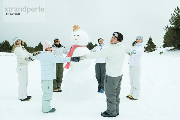 Junge Freunde stehen im Kreis um den Schneemann  halten sich an den Händen und lächeln in die Kamera.