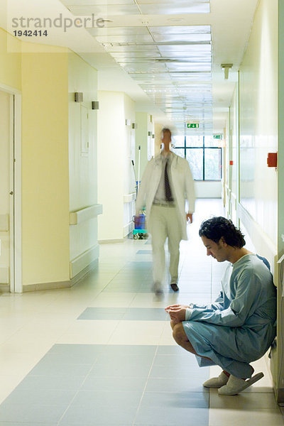Männlicher Patient hockt im Krankenhausflur  schaut nach unten  Arzt läuft im Hintergrund