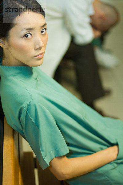 Ärztin sitzt mit der Hand in der Tasche  schaut in die Kamera  Ärztin mit dem Kopf nach unten im Hintergrund.
