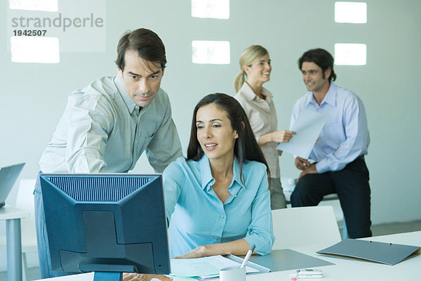 Geschäftsmann und Geschäftsfrau im Büro  Blick auf den Computer  Mitarbeiter im Hintergrund