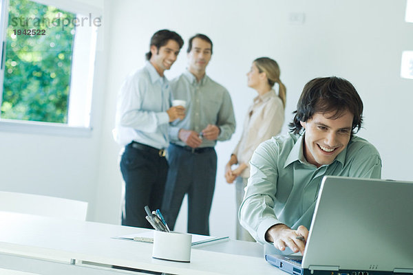 Geschäftsmann im Büro  mit Laptop  lächelnd  Mitarbeiter im Hintergrund