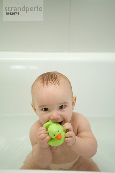 Nacktes Baby sitzend in der Badewanne  beißende Badeente  Blick in die Kamera