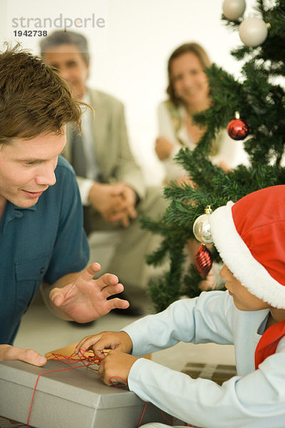 Vater und Sohn eröffnen Weihnachtsgeschenk vor dem Weihnachtsbaum  Junge mit Weihnachtsmütze